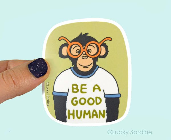 Sticker of a monkey wearing glasses with 'Be a Good Human' text, held by a hand against a light blue background.