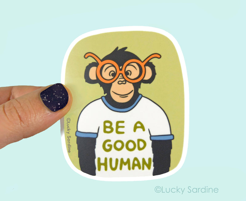 Sticker of a monkey wearing glasses with 'Be a Good Human' text, held by a hand against a light blue background.