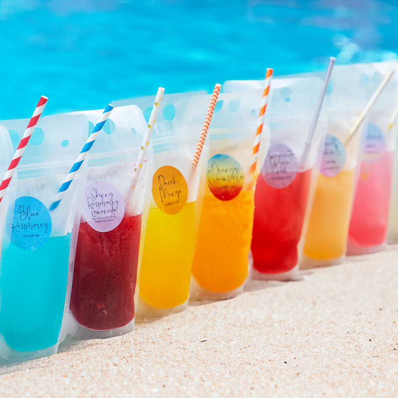 Colorful drinks in bags with straws on a sandy beach by a pool.
