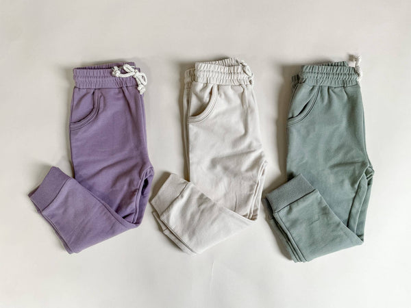 A set of three pairs of baby pants in different colors: purple, white, and gray, all with drawstrings and pockets.