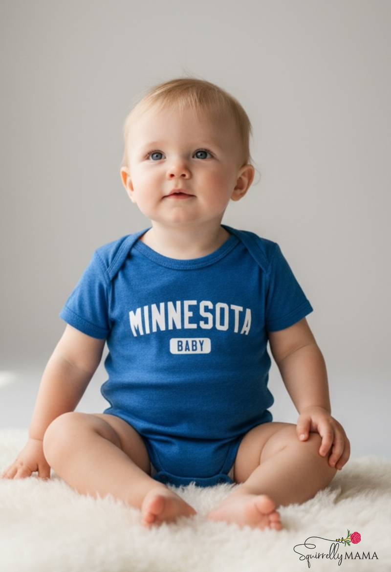 Baby wearing a blue 'MINNESOTA BABY' onesie sitting on a white surface.