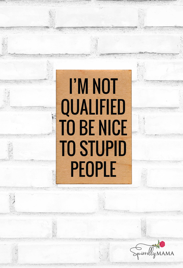 Wooden magnet with text 'I'm not qualified to be nice to stupid people' on a white brick wall.