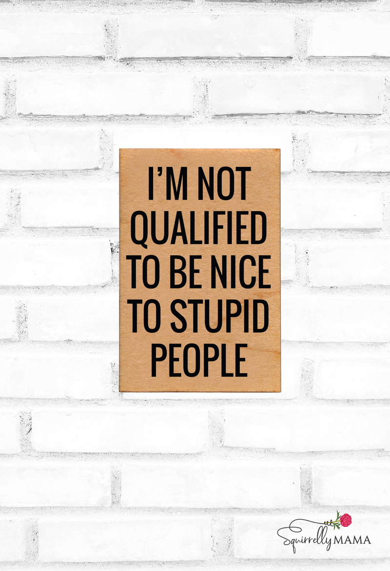 Wooden magnet with text 'I'm not qualified to be nice to stupid people' on a white brick wall.