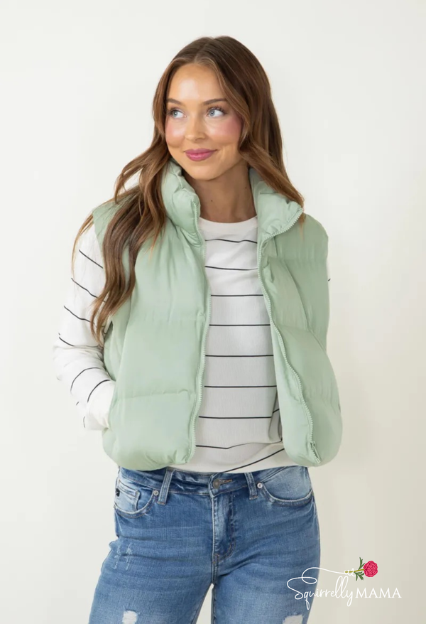 Woman wearing a light green puffer vest over a striped shirt and jeans on a white background.