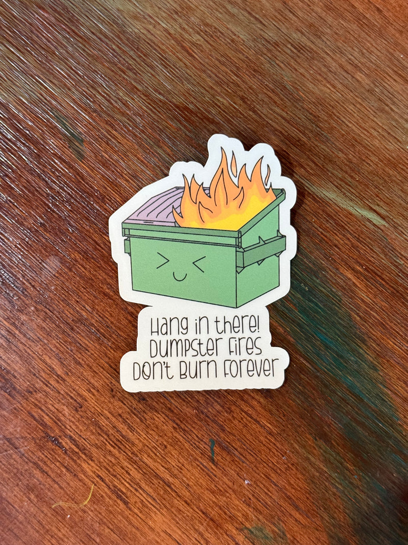 Sticker with a cartoon dumpster on fire and text 'Hang in there! Dumpster fires don't burn forever' on a wooden surface.