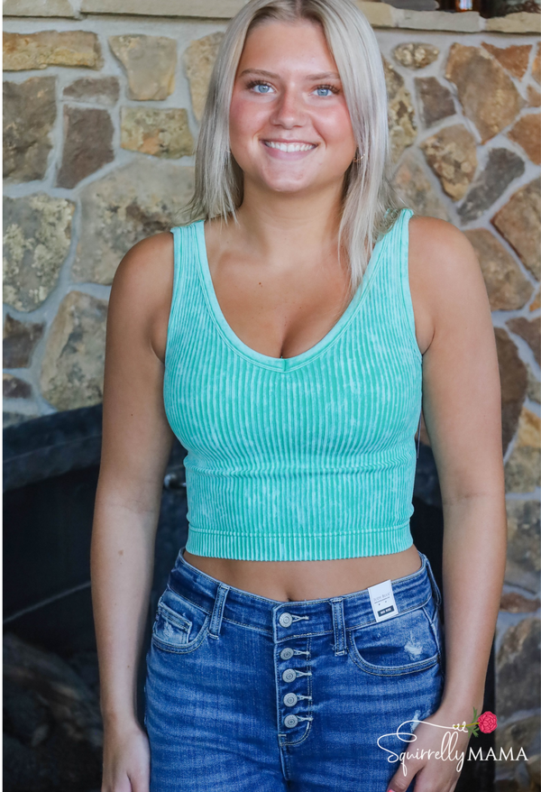 Woman wearing a teal ribbed tank top and blue jeans standing in front of a stone wall.