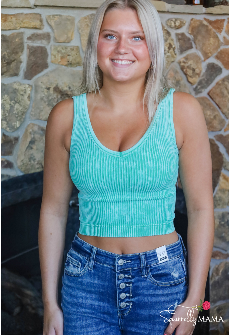 Woman wearing a teal ribbed tank top and blue jeans standing in front of a stone wall.