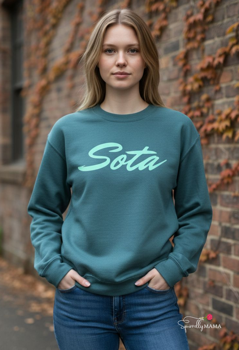 Person wearing a teal sweatshirt with 'Sota' printed on it, standing against a brick wall.